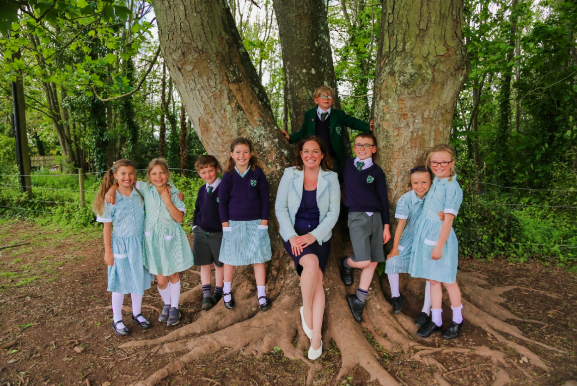 A group of school students standing around a teacher by a tree smiling