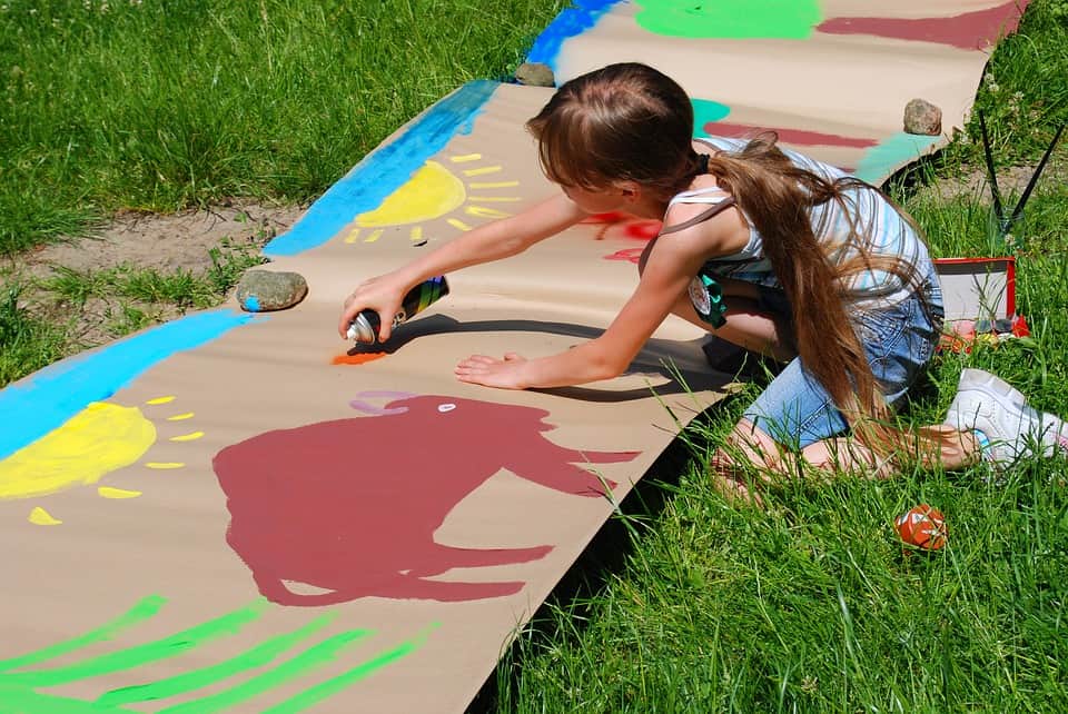 A young girl using spray paint to craft a large painting on cardboard