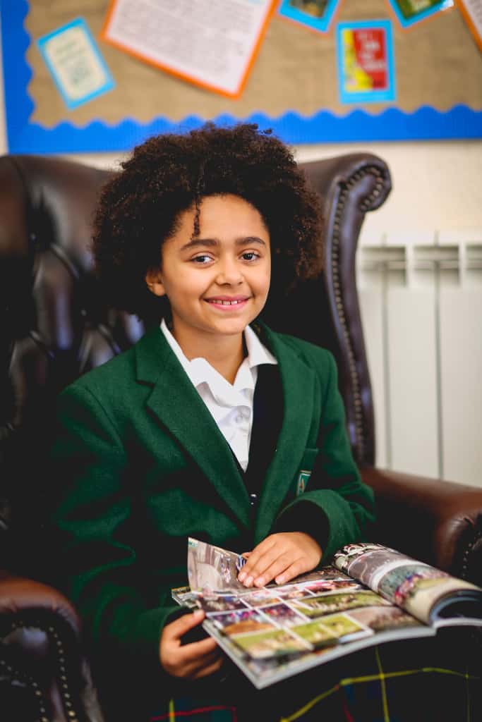 A girl sitting on a chair in school with a magazine on her lap, smiling
